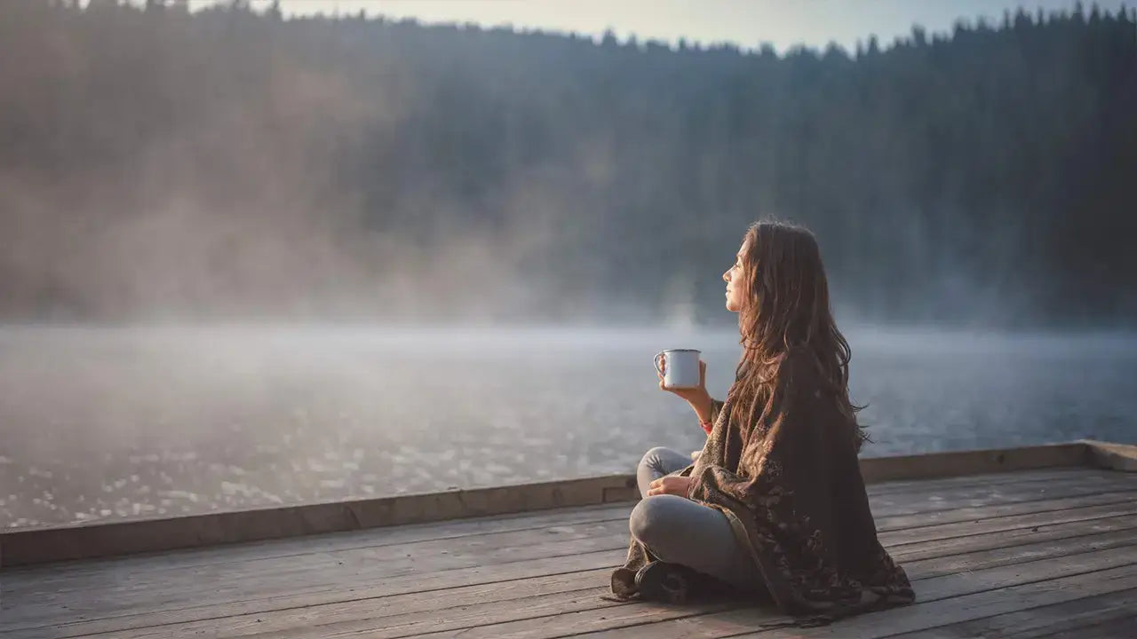 Eine Frau sitzt mit einer Tasse Tee auf einem Steg am See - Wenn man gerade keine malerischen Ort bewohnt, an dem Entspannung herscht, kann die Akupressurmatte helfen